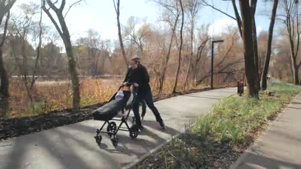 Jonge familie wandelen in de herfst met een baby in een kinderwagen op rolschaatsen in een zonnig stadspark. Papa rijdt op een ander spoor en haalt mam in. Langzame beweging. — Stockvideo