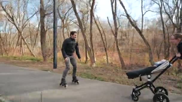 Şehir parkında bebek arabası ve paten kullanan mutlu ebeveynler. Peder paten kayıyor. Annem bebek arabasını ileri itiyor, babam da onu yakalıyor. Yavaş çekim. — Stok video