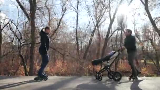 Deportiva joven familia de patinadores con un bebé en un cochecito en un acogedor parque de otoño. Mamá cabalga hacia atrás y entretiene al niño. Movimiento lento . — Vídeos de Stock