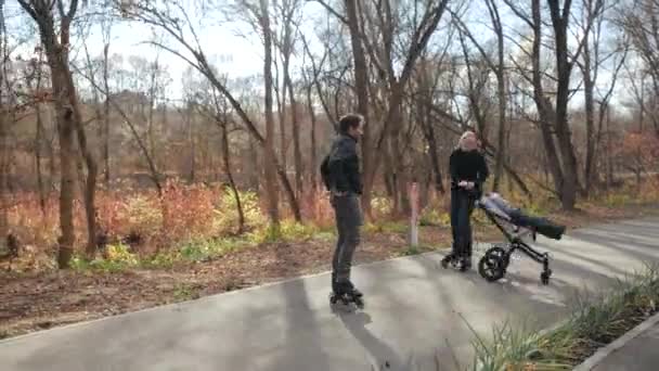 De jeunes parents en rollers marchent à l'automne avec un enfant dans une poussette dans un parc urbain ensoleillé. Maman déballe la poussette et roule avec papa de l'autre côté. Mouvement lent . — Video