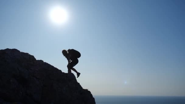 Sporty woman climbs to the top of a high cliff with a backpack and looks at a pretty view with the sun and the falling rays of the sun on the sea in slow motion, side view. Active leisure outdoor. — ストック動画