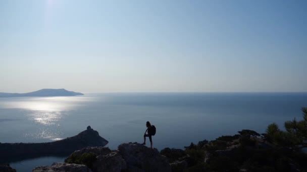 Woman with a backpack admires a beautiful wide view of the sea under the sun and waves hands, standing on a very high rock, shooting from above. Sport tourism and active life concept in slow motion. — Stock Video