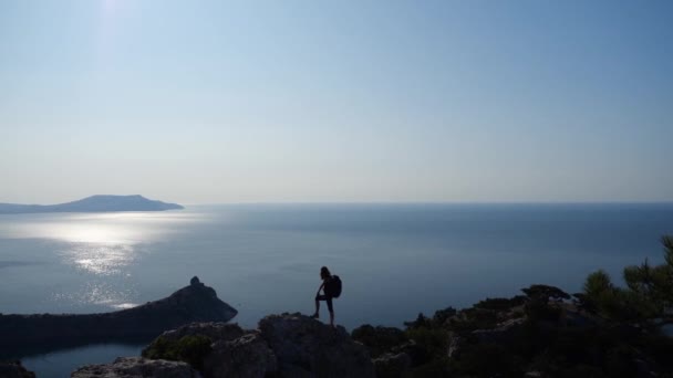 Γυναίκα με σακίδιο θαυμάζει μια όμορφη θέα της θάλασσας κάτω από τον ήλιο και τα χέρια κύματα, στέκεται σε ένα πολύ ψηλό βράχο, γυρίσματα από ψηλά. Αθλητισμός τουρισμός και ενεργός ζωή έννοια σε αργή κίνηση. — Αρχείο Βίντεο