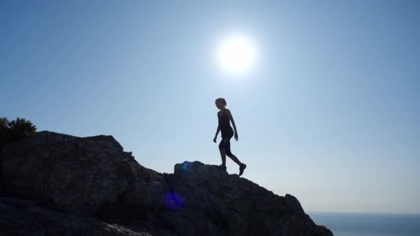 Atletische jonge vrouw klimt naar de top van de klif en steekt haar handen op in plezier. Zijaanzicht van een sportief mooi meisje genieten van schoonheid met uitzicht op de zee en de felle zon in slow motion. — Stockvideo