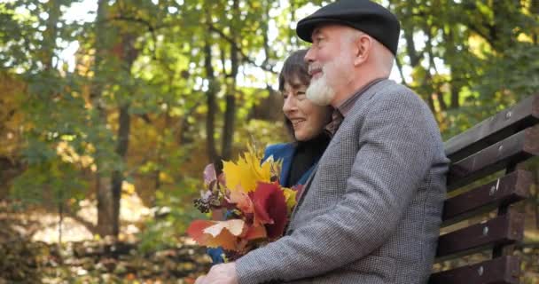 Vue latérale de l'homme et de la femme âgés passent du temps ensemble à l'extérieur, assis sur un banc et bavarder dans un parc ensoleillé de la ville confortable. La femme caresse son mari avec un bouquet de feuilles d'automne au ralenti . — Video