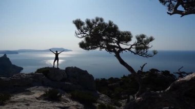 Atletik genç kadın denizin üzerindeki yüksek bir uçurumun tepesinde duruyor ve denizin, dağların ve kayaların üzerinde parlayan göz kamaştırıcı güneşine bakıyor. Seyahat ve turizm fikri.