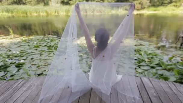 The bride sits on a small bridge over the water and puts a veil on her head, back view. A girl sits and rests on a wooden pontoon near a beautiful colorful autumn forest mirrored in a mountain lake. — Stock Video