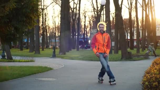 Hombre barbudo fresco con abrigo naranja y pantalones vaqueros baila en patines en otoño en un parque central de la ciudad sobre un fondo de árboles. Deportes activos y estilo de vida saludable. Vista lateral en cámara lenta . — Vídeos de Stock