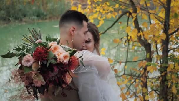 Gros plan portrait de Mariée et marié embrassant près d'un lac dans la forêt d'automne parmi les arbres d'automne colorés. Jeune attrayant heureux aimer les jeunes mariés dans un parc au ralenti . — Video