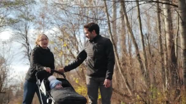 Front shot of a happy young family roller skating in a cozy city park in autumn in slow motion. Husband and wife are carrying a carriage with a baby, sliding on the asphalt. Sports family vacation. — Stock Video
