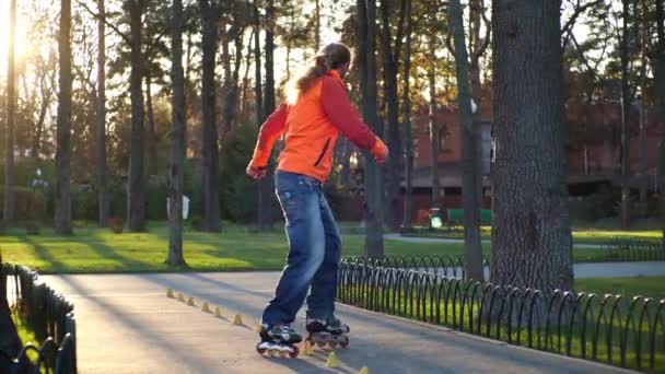 El hombre en los rodillos realiza varias vueltas y paseos entre los conos de entrenamiento, cruzando sus piernas. Entrenamiento profesional de patinaje sobre ruedas en un parque de la ciudad en otoño con clima soleado. Primeros planos en cámara lenta . — Vídeo de stock