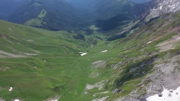 Terugvliegend over een bergdal in de bergen van Adygea op zomerdag. Groene bomen en struiken tussen rotsen en sneeuwvelden. Wilde natuur van bovenaf gezien. — Stockvideo