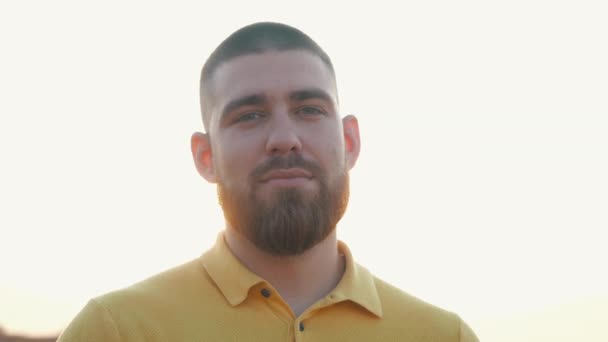 Close-up view of the face of a short-haired bearded man in a yellow polo shirt standing on the field in autumn. Nice face of a man in front view. Portrait of a young beard european man in slow motion. — Stock Video