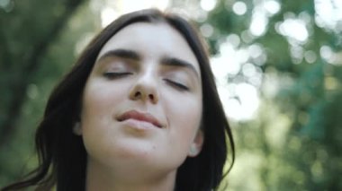 Güzel bir gülümsemesi olan güzel bir kadın ormanda kameraya poz veriyor. Beyaz gömlekli güzel kız güzel bir fotoğraf için gülümsüyor. Kadın gözlerini kapattı ve parkın temiz havasını ve doğasını seviyor..