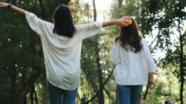Beyaz tişörtlü, uzun saçlı, dans eden ve güzel fotoğraflar için ağır çekimde zıplayan iki çekici kızın sırtından. Güneşli bir bahar gününde fotoğraf çekimi yapan bir çift güzel genç kadın..