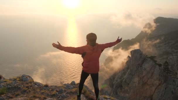 Disparo desde la espalda cerca de un hombre barbudo en un chándal, que está de pie en el borde de una alta montaña, se levanta y levanta los brazos aparte de disfrutar de la hermosa vista del mar y la puesta de sol . — Vídeos de Stock