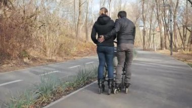 Neşeli ve hareketli bir baba ve anne, bebek arabasıyla ve bebek arabasıyla park yolu boyunca yürüdüler. Sıcak, sıcak bir parkta güneşli bir günde spor ebeveynleri asfaltta kayıyorlar..