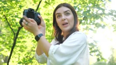Şehir parkındaki parlak bahar güneşinin karşısında arkadaşına kameradan çekilen fotoğrafları gösteren güzel bir genç kızın yakın görüntüsü. Profesyonel bir fotoğrafçı kız, çekilen fotoğraflara bakar..
