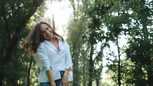 Servizio fotografico di primavera per una ragazza carina in camicia bianca e jeans. Giovane donna allegra con i capelli lunghi e la manicure bianca sta ballando alla telecamera tra gli alberi del parco nella giornata di sole. Video per memoria . — Video Stock