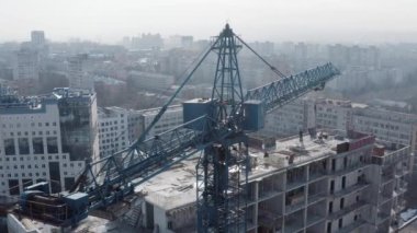 Circular top shot of the crane and the process of building a new multi-storey building in the winter season against the background of residential buildings. Workers on the roof are building a house.