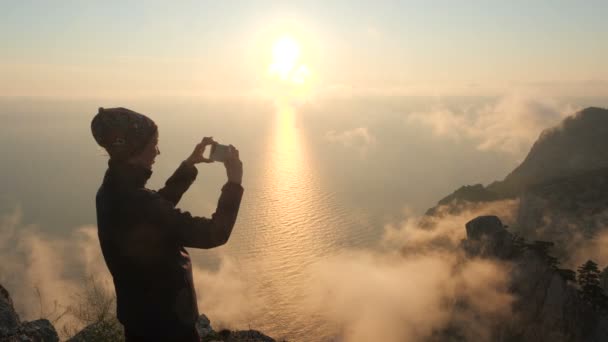 Close-up shooting of a sporty tourist woman wearing a bandana standing on the edge of a high mountain, who photographs a beautiful sunset and cloud smoke and steam over the sea on her smartphone phone — ストック動画