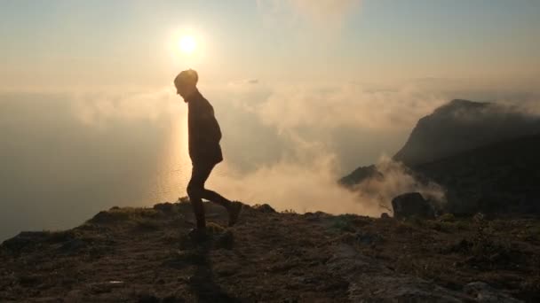 Chica en una capucha camina cerca del borde de una montaña sobre el mar en el contexto de una hermosa puesta de sol en otoño. Persona camina cerca de un acantilado de una montaña, vapor de nubes es visible desde abajo . — Vídeos de Stock