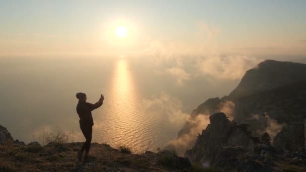 Letecký záběr v blízkosti mladé turistické dívky, která stojí na okraji vysokého útesu a bere různé selfie z jejího smartphonu na pozadí nádherného západu slunce na podzim. — Stock video