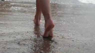 Shooting behind the beautiful feet of a young girl walking along the small foamy waves of the sea along the sandy shore with small pebbles. Slender female legs gracefully walk along the ocean.