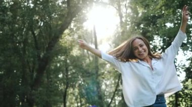 Front view of a young pretty girl in a white shirt and long hair who is looking at the camera and bouncing for a nice fun shot in a warm cozy park among green trees and the shining sun between them.