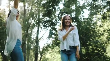 Bir çift mutlu güzel kız arkadaş, şehrin sıcacık bir parkında, manzaranın önündeki ağaçların arasında eğlenceli bir fotoğraf çekimi yapıyorlar. Güneşli bir yaz gününde iki çekici kadın zıplıyor ve kameraya gülümsüyor..