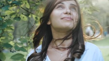 Beautiful lovely girl with green eyes and long black hair smiles into the lens and beckons. Photoshoot for a pretty young woman in a city park against a background of trees and a bright shining sun.