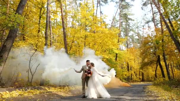Gelukkig liefdevolle pasgetrouwden tussen gekleurde bomen in een park met brandende rookbommen. Langzame beweging. Jonge aantrekkelijke bruid en bruidegom lopen langs een weg in de herfst bos. — Stockvideo