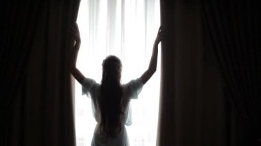 A young, long-haired woman runs her hands over the curtains in the bedroom, shielding herself from the morning light that comes in through the window. Beautiful girl opens the curtain on the window