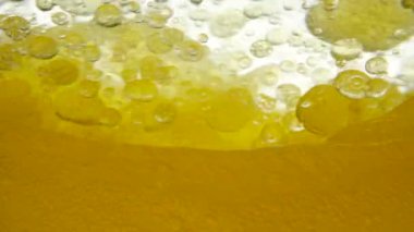 Mixing black caraway seed oil and linseed oil as a result of which air bubbles and a droplet structure are created, close-up. The heterogeneous phase of the golden liquid on a moving white background.