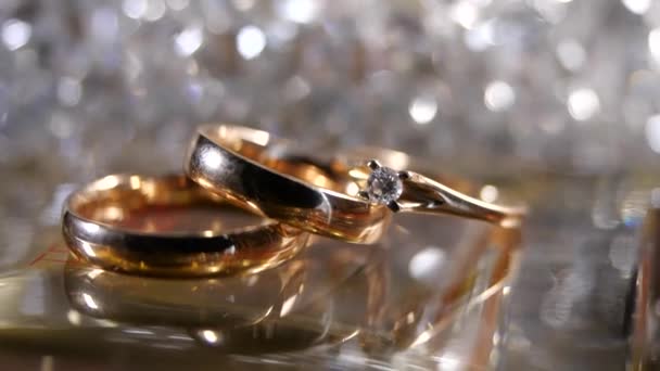 Tre bellissimi anelli d'oro un anello di fidanzamento di diamanti e un anello di fidanzamento si trovano su uno sfondo scintillante e scintillante. Regali e romanticismo, arredamento, sposa e sposo, luce cangiante e abbagliamento — Video Stock