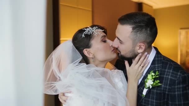 Vista da vicino di una coppia felice che si bacia in una camera di lusso di un hotel costoso. Uno sposo barbuto in abito abbraccia e bacia la sua amata sposa in un abito bianco chic. L'inizio della luna di miele . — Video Stock