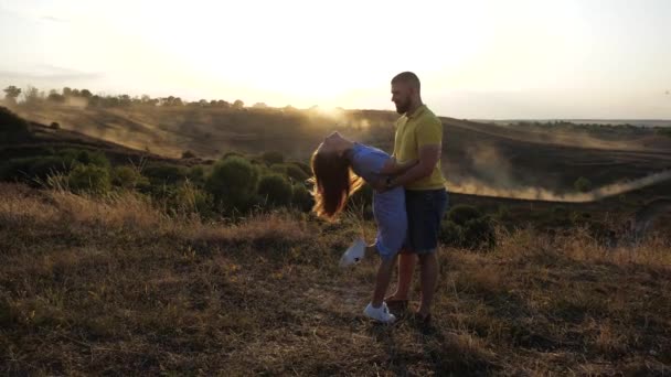 Romantiskt datum för ett ungt par ute på fältet i varmt väder. Skäggig kille lutar flickan som håller henne i ryggen. Två älskare tillbringar tid tillsammans på ängen mot bakgrund av gryningen. — Stockvideo