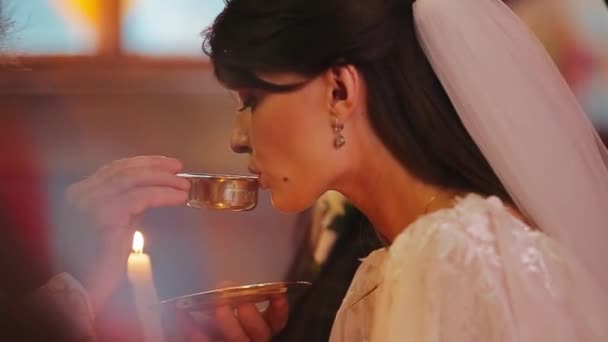 Hermosa novia en lujoso vestido blanco bebe agua sagrada en copa dorada en las manos del sacerdote. La tradición cristiana de la boda de la iglesia en Ucrania. La costumbre europea para los creyentes enamorados en la ceremonia . — Vídeos de Stock