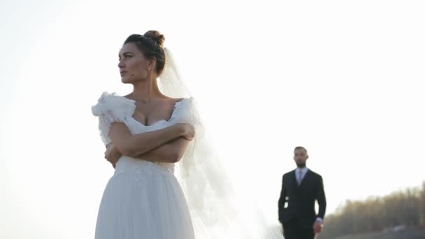 Beautiful bride in a chic white dress is standing in field, looking to the side and waiting for her groom, who is slowly approaching from behind, focusing on the girl. Newlyweds walk in the fresh air. — Stockvideo