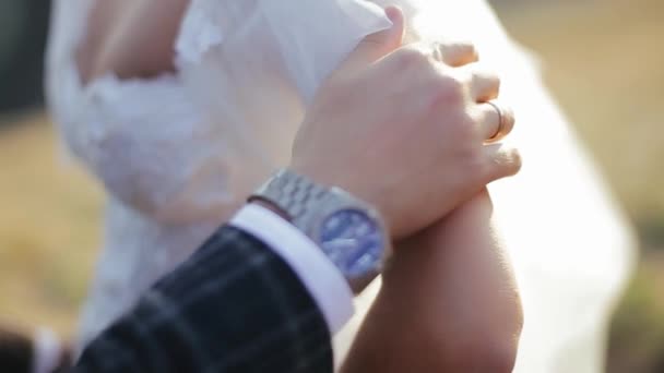 Young bearded groom strokes his bride by the hand on the nature, focusing on the girl, close view. Pretty bride in a white dress is enjoying a romantic evening with her beloved in a field under sun. — Stok video