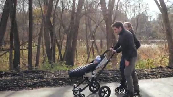 Gelukkige jonge familie wandelen op rolschaatsen in het park. Mam en pap rolschaatsen met een baby in een kinderwagen. Veilig actief type recreatie en buitensporten in het park op herfstdag in de steeg. — Stockvideo