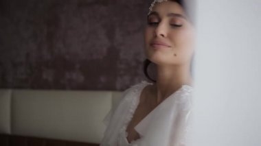 Beautiful millennial bride with a wreath on her head and in a white wedding dress holds a bouquet of flowers in the bedroom in the morning in the hotel room, close shot from behind the curtains.