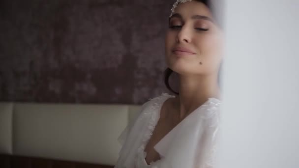 Beautiful millennial bride with a wreath on her head and in a white wedding dress holds a bouquet of flowers in the bedroom in the morning in the hotel room, close shot from behind the curtains. — ストック動画