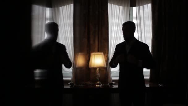 Silhouette eines stilvollen reichen Mannes, der in einem dunklen Raum am Fenster steht und eine schöne Lampe einschaltet und seinen teuren Anzug in einem teuren Hotelzimmer richtet. Vorbereitung auf ein Geschäftstreffen. — Stockvideo