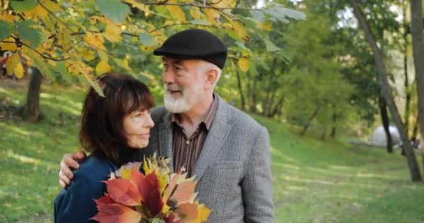 Щасливий старший чоловік і жінка з букетом листя, що обнімається в теплому затишному парку в сонячну погоду. Літня пара відпочиває навколо осінніх дерев. старший громадянин обіймає, заспокоює і втішає свою дружину — стокове відео