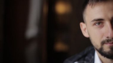 Sabahleyin genç, beyaz sakallı yakışıklı damadın yüzünün yarısını pahalı bir otel odasının karanlık duvarlarına yakın çekim. Yakışıklı Avrupalı adam kameraya bakıyor..