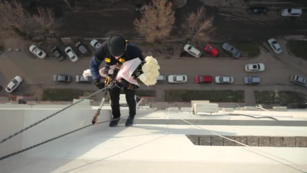 Hombre escalador industrial que cuelga de la cuerda de seguridad, descendiendo a lo largo de la pared de la casa con ramo de rosas blancas elegantes. Entrega de regalos inusual. Sorpresa peligrosa, regalo creativo inusual para su amada . — Vídeos de Stock