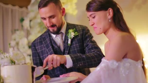 Šťastný kavkazský svatební pár vousatý ženich a krásná tisíciletá nevěsta krájet kus lahodný a chutný bílý dort pro hosty v hodovní síni na party na pozadí oltáře. — Stock video