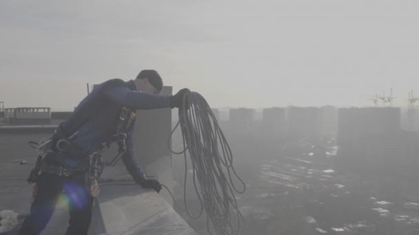 Чоловічий альпініст у спеціальній формі стоїть на даху і скидає кабель, на який він спуститься ззовні, з близьким видом на місто в повільному русі. Небезпечна і екстремальна робота для чоловіків . — стокове відео