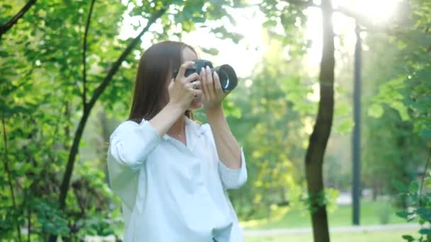 长发的年轻漂亮姑娘用专业相机拍照。迷人的女人穿着白衬衫，在春天里温暖的阳光灿烂的日子里，拍下了一个舒适的城市公园的自然美景. — 图库视频影像